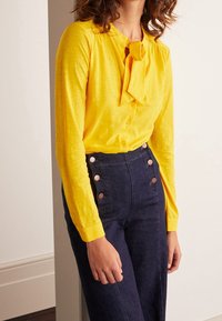 Blusa amarilla de mangas largas con detalle de lazo, combinada con pantalones de mezclilla oscuros de tiro alto con botones de color cobre.