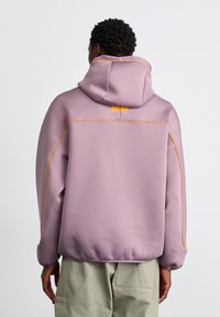 Hoodie i ljus lila tyg med gula sömmar. Har en avslappnad passform, känguruficka och ribbade muddar och fåll.