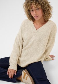 Lys beige v-hals sweater i et blødt, struktureret strik. Førstegangsparret med mørke jeans og beige sneakers med brune detaljer.
