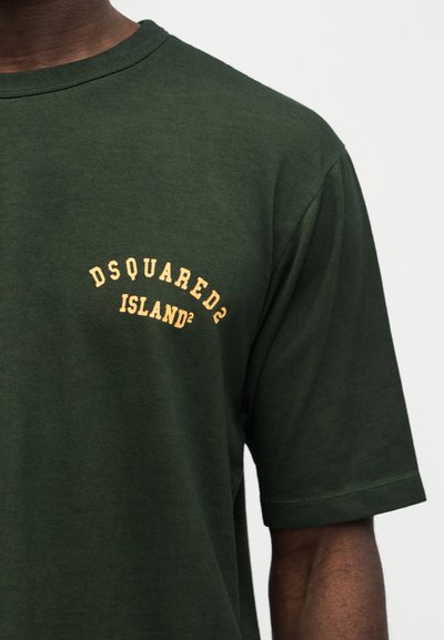 Σκούρο πράσινο κοντομάνικο μπλουζάκι με κίτρινο κείμενο "DSQUARED2 ISLAND²" στο αριστερό στήθος, φορεμένο από άτομο με σκούρο τόνο δέρματος.