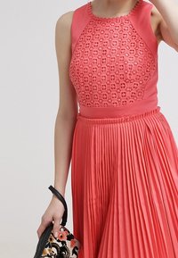 Koronkowa sukienka o koralowym kolorze z wzorzystym torsem i plisowanym dołem. Posiada wysoki dekolt i teksturowany wzór. Ręcznie robiona czarna kopertówka w kwiaty.