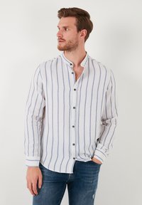 Camisa blanca de manga larga con rayas verticales azules, cuello abotonado y botones en contraste. Llevada con unos vaqueros oscuros informales.