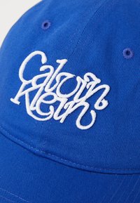 Blauwe katoenen baseballpet met een witte geborduurde "Calvin Klein" logo, textuurstof en gestikte details op de klep.