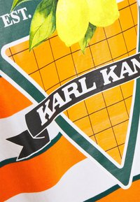 Oranžovo-bíle pruhovaná látka s zeleno-žlutým trojúhelníkovým logem s limonádami a textem "KARL KANI" na mřížkovém pozadí.