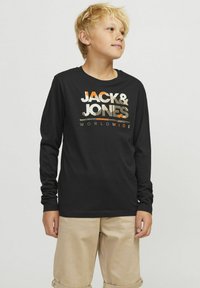 Crna majica dugih rukava s logom "JACK & JONES" u metalnim nijansama sprijeda; uparen s svijetlim bež kratkim hlačama. Mekani materijal i ležeran kroj.