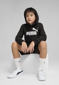 Crna dukserica s bijelim "PUMA" logotipom, odgovarajuće crne kratke hlace, bijele čarape s logotipom i bijele tenisice s crnim akcentima. Opusten položaj.