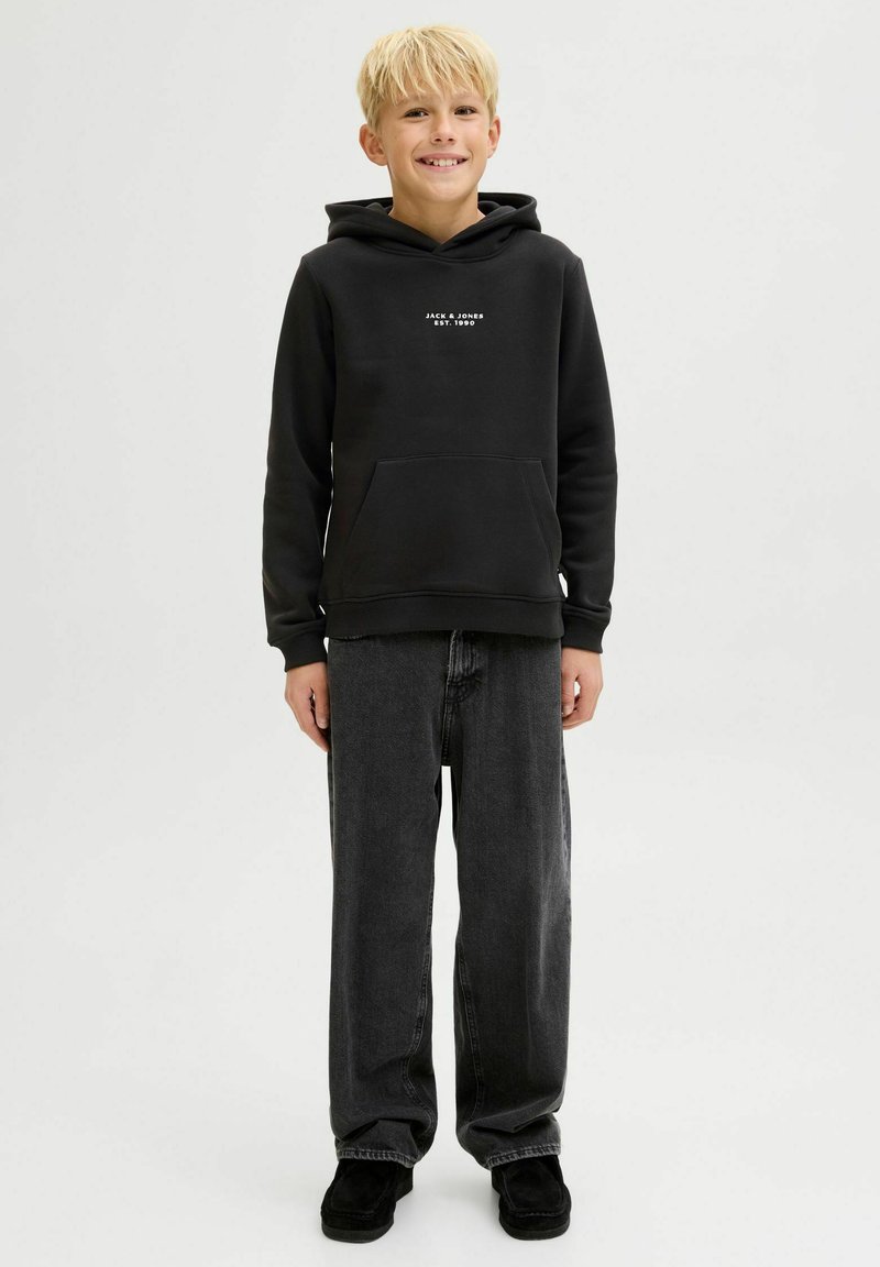 Sudadera negra con bolsillo canguro y logo blanco en el pecho, combinada con jeans sueltos de color gris oscuro y zapatos negros.