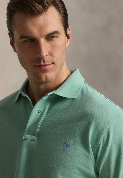 Νεαρός άνδρας με κοντά μαλλιά που κοιτάζει προς τα κάτω, φοράει ανοιχτοπράσινο πόλο με ένα μικρό μπλε κεντητό λογότυπο στο στήθος.