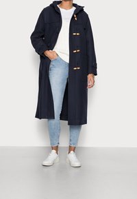 Granatowy płaszcz z kapturem, z zapięciem na toggle i kieszeniami z przodu, nałożony na biały sweter, w połączeniu z jasnoniebieskimi dżinsami i białymi trampkami.