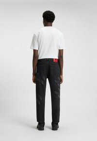 Black denim jeans with a subtle diagonal pattern, tag on the back, worn with a plain white t-shirt, paired with black shoes.