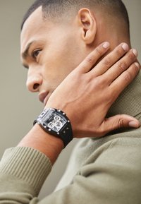 Montre rectangulaire noire avec des cadrans argentés et des accents métalliques sur un bracelet noir. Portée sur une main à la peau claire contre un pull en laine vert texturé.