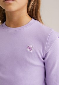 Šviesiai violetinė ilgomis rankovėmis, išmegzta megztinio marškinėliai su apvalia iškirpte. Kairėje krūtinės pusėje yra mažytis siuvinėtas gėlės atspalviai violetinės spalvos.