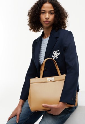 Kvinde med krøllet hår iført en marineblå blazer og en lyseblå sweater holder en tanfarvet håndtaske, mens hun sidder mod en hvid baggrund.