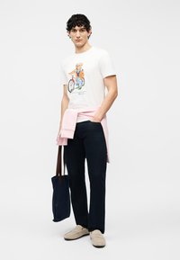 Άνδρας που φοράει λευκό μπλουζάκι με σχέδιο, μαύρο παντελόνι, μπεζ παπούτσια, ροζ πουλόβερ δεμένο γύρω από τη μέση, κρατώντας σκούρη μπλε τσάντα tote, στέκεται μπροστά σε λευκό φόντο.