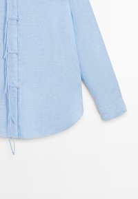 Lichtblauw overhemd met lange mouwen en een textuur in de stof, voorzien van knoop- en lussluitingen aan de voorzijde aan de linkerkant.