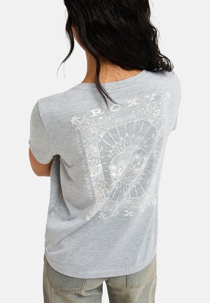 Person mit dunklen Haaren, die ein hellgraues T-Shirt mit weißem Grafikdesign und Text auf der Rückseite trägt und mit verschränkten Armen steht.