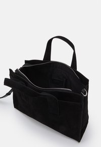 Zwarte suède tas met dubbele handvatten aan de bovenkant, ritssluiting en een gestructureerde buitenkant. Het interieur heeft voering en een label. Minimalistisch ontwerp.