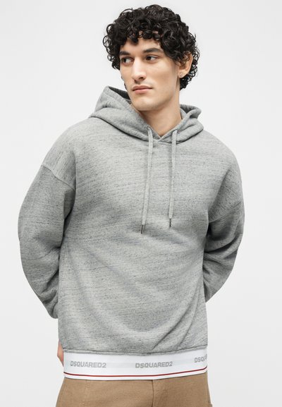 Giovane uomo con capelli ricci e scuri che indossa una felpa con cappuccio grigio chiaro con coulisse e pantaloni beige, che guarda verso sinistra contro uno sfondo semplice.