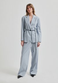 Svetlo modra obleka po meri z širokimi hlačami in prilegajočim blejzerjem. Vključuje črn pas kot dodatek in črne špičaste čevlje. Gladka tekstura.