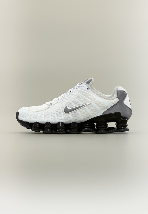 Bílá a šedá teniska Nike s černou segmentovanou podrážkou, síťovaným svrškem a tkaničkami, zobrazena z boku na neutrálním pozadí.