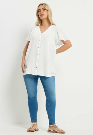 Vaaleatukkainen nainen seisoo käsi lanteilla, pukeutuneena valkoiseen napitettuun V-aukkoinen paitaan, sinisiin kapeisiin farkkuihin ja beigenvärisiin remmisandaaleihin.