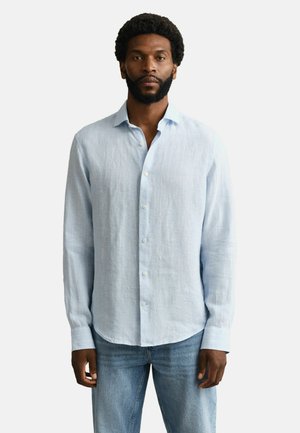 Man met kort krullend haar en baard staat frontaal en draagt een lichtblauw overhemd met knopen en een blauwe spijkerbroek tegen een witte achtergrond.