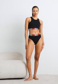 Černý bikiny set s crop topem, který má červenobílý pás s logem "DIESEL". Hladký materiál, vysoký střih nohaviček, minimalistický styl.