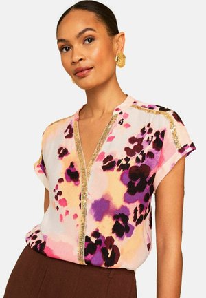 Žena so stiahnutými vlasmi, ktorá nosí zlaté náušnice a farebnú kvetinovú blúzku so zlatým lemom, v kombinácii s hnedými nohavicami.