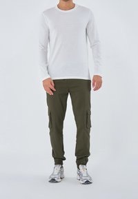 Bílá dlouhá košile s kulatým výstřihem, v kombinaci s olivově zelenými cargo kalhotami s bočními kapsami a elastickými manžetami, doplněná šedými teniskami.