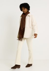 Chaqueta de borreguillo crema de gran tamaño con cuello, bufanda marrón con flecos, pantalones crema y zapatos de tacón cuadrado de ante marrón.