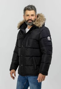 Giubbotto puffa nero con cappuccio foderato in pelliccia, dotato di zip frontale, chiusura a bottoni e tasche laterali, indossato sopra una camicia bianca.