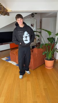 Melnā kapuce ar baltu grafisku apdruku, vaļīgas melnas bikses un sudraba sporta apavi. Telpās ir redzams augs un oranžs kordurojas dīvāns.