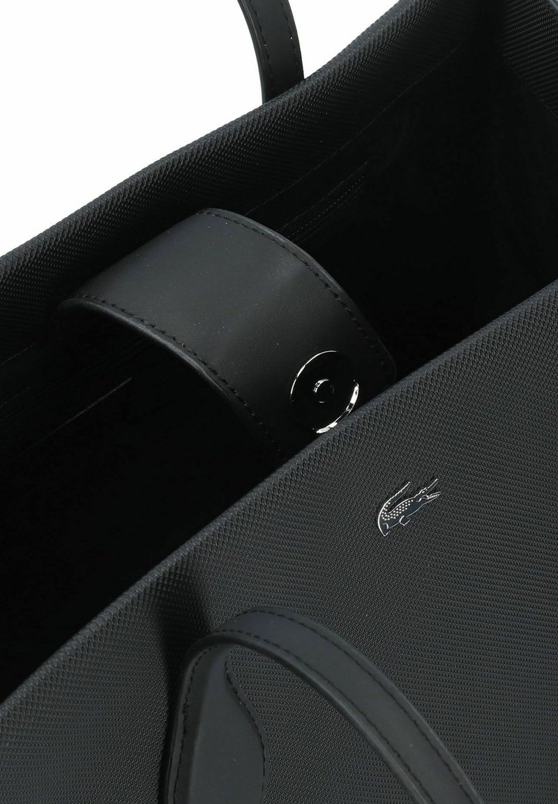 Sac fourre-tout noir présentant un extérieur en tissu texturé, une poignée en cuir et des accessoires en métal argenté avec un accent logo. À l'intérieur, un revêtement lisse et sombre.