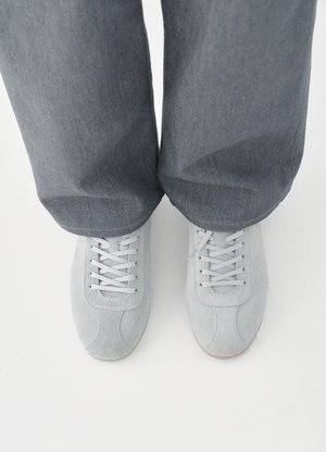 Αθλητικά παπούτσια από ανοιχτό γκρι σουέντ με στρογγυλεμένη μύτη, σχέδιο με κορδόνια και διακριτικές ραφές, συνδυασμένα με σκούρο γκρι παντελόνι από τζιν.