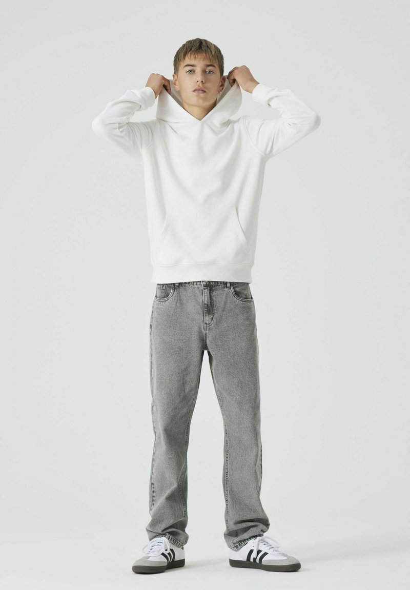 Sweatshirt blanc en coton avec une poche kangourou, associé à un jean en denim gris clair. Les chaussures comprennent des baskets noires et blanches avec des rayures.