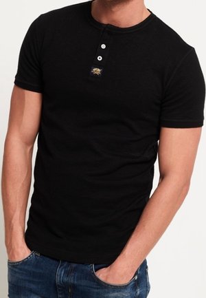 Czarny t-shirt z krótkim rękawem, dekoltem typu crew neck i patką na trzy guziki, z małą naszywką z logo nad lewą piersią. Casualowy krój.