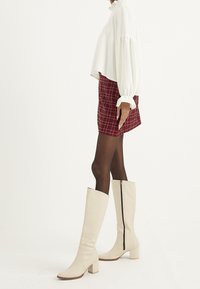 Blusa blanca corta con mangas abullonadas, falda mini de cuadros rojos y botas beiges hasta la rodilla con cremallera lateral y tacón cuadrado.