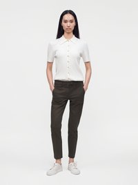 Femme debout, les mains dans les poches, portant une chemise blanche à manches courtes boutonnée, un pantalon slim foncé et des baskets blanches sur un fond uni.