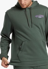 Felpa verde scuro con tasca a marsupio frontale, cappuccio regolabile con lacci neri e oro, e logo viola sul petto.