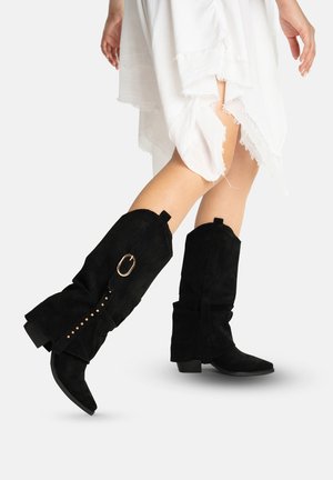 Beine mit schwarzen Wildlederstiefeln bis zur Mitte der Wade, mit gestapelten Absätzen und goldenen Schnallendetails, kombiniert mit einem weißen asymmetrischen Rock mit ausgefranstem Saum.