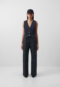 Tmavě modrá džínová vesta s nízkým V výstřihem a zapínáním na knoflíky, spárovaná s černými prošívanými kalhotami s logem a stahovací šňůrkou. Černá kšiltovka a elegantní obuv.