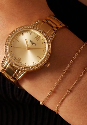 Gold watch with a round face, crystal-accented bezel, and gold link bracelet. Minimalistic dial with Roman numerals and brand logo. Twin gold chain bracelet.