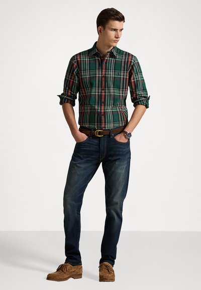 Camisa de cuadros verde y con botones, con las mangas arremangadas, combinada con jeans oscuros y zapatos de ante marrones. Incluye un cinturón marrón y un reloj de pulsera.