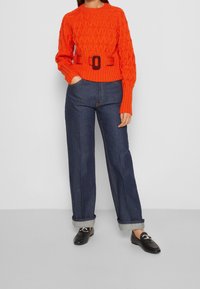 Pull en crochet orange vif avec une ceinture ajustée, porté avec un jean en denim bleu foncé à jambes larges et ourlets retroussés. Des mocassins noirs complètent le look.