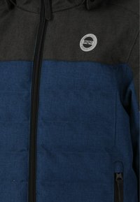 Blå og sort lynlåsjakke med struktureret stof, en rund logo-patch på brystet og sidelommer med sorte lynlåse.