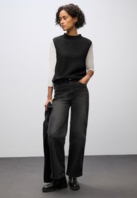 Top nero senza maniche sopra una camicia bianca a maniche lunghe, abbinato a jeans neri a vita alta con gamba larga e stivaletti neri. Design semplice.