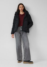 Schwarze Pufferjacke mit Kapuze, über einem burgunderfarbenen Pullover getragen, kombiniert mit hochgeschnittenen grauen weit geschnittenen Jeans und schwarzen Plateaustiefeln.