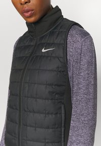 Černá zateplená vesta s prošívanou texturou, vysokým límcem a logem Nike. Má boční panely z tmavě šedé látky s jemnou texturou.