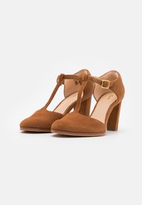 Chaussures à talons hauts en suède marron avec une bride T, fermeture à boucle, bout rond et talons épais sur fond blanc.