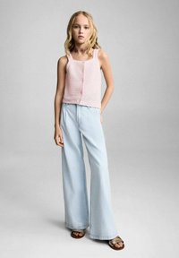 Svetlo roza pletena majica brez rokavov z teksturiranim vzorcem, v kombinaciji s svetlo modrimi širokimi hlačami in rjavimi sandali. Preprost, sproščen dizajn.